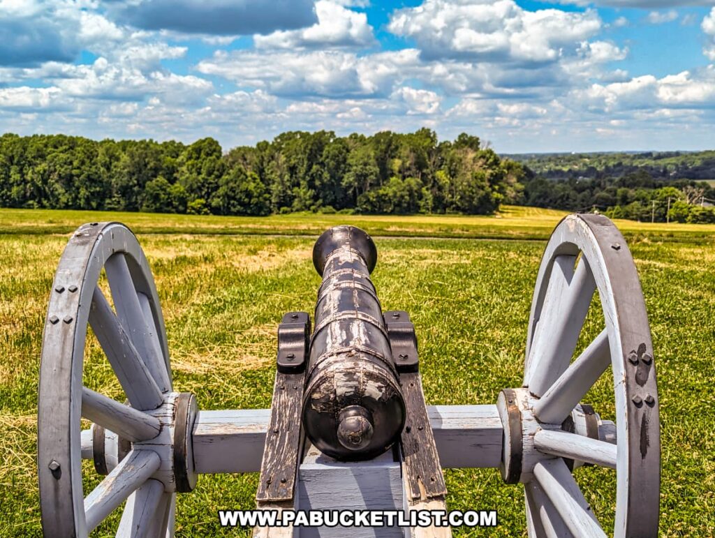 A historic cannon at Brandywine Battlefield Park in Chester County, Pennsylvania, is positioned on a grassy field, facing a dense tree line in the distance. The view from behind the cannon barrel emphasizes its strategic placement during the Battle of the Brandywine. The sky is bright blue with scattered white clouds, highlighting the lush green landscape. This scene evokes the historical context of the largest and longest single-day land battle of the American Revolution, emphasizing the park's role in preserving and interpreting this significant event.