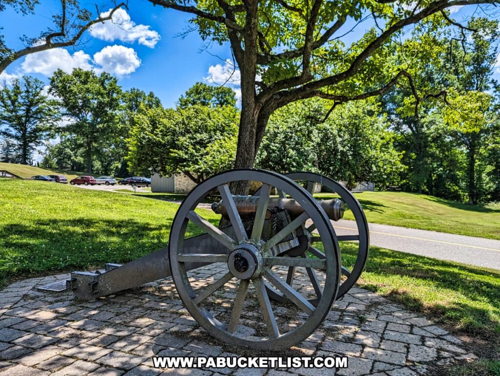 A historic cannon is displayed on a paved area under a tree at Brandywine Battlefield Park in Chester County, Pennsylvania. The cannon is positioned near the visitor center, visible in the background along with a parking area and other park amenities. The scene is set on a sunny day with a bright blue sky and scattered white clouds, highlighting the lush green grass and foliage. This setting underscores the park's historical significance as the site of the largest and longest single-day land battle of the American Revolution.