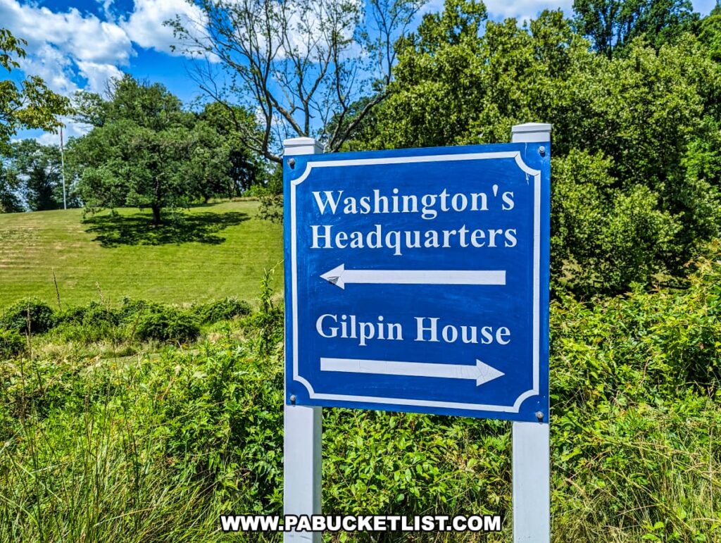A blue directional sign at Brandywine Battlefield Park in Chester County, Pennsylvania, provides guidance to visitors. The sign indicates the direction to Washington's Headquarters with an arrow pointing left and the Gilpin House with an arrow pointing right. The sign is set against a backdrop of green foliage and trees under a bright blue sky with scattered white clouds. The well-maintained grassy area surrounding the sign highlights the park's natural beauty and historical significance, commemorating the largest and longest single-day land battle of the American Revolution.