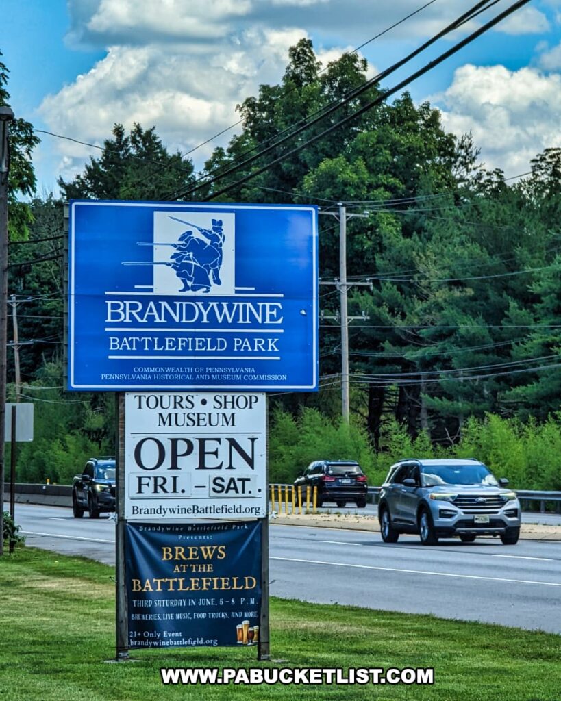 A blue and white entrance sign for Brandywine Battlefield Park in Chester County, Pennsylvania, is prominently displayed along a busy road. The sign includes the park's name and logo, which features a silhouette of Revolutionary War soldiers. Below the main sign, additional information indicates that the park's tours, shop, and museum are open on Fridays and Saturdays. Another banner promotes the "Brews at the Battlefield" event, held on the third Saturday in June, featuring breweries, live music, food trucks, and more. Cars are visible on the road behind the sign, and the surrounding area is lush with trees and greenery, emphasizing the park's accessibility and community engagement.