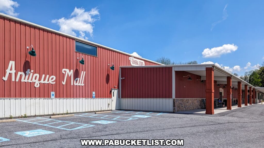The exterior of Cackleberry Farm Antique Mall in Lancaster County, PA, featuring a large red building with white accents and a sign reading "Antique Mall." The entrance area is covered, providing a shaded walkway, and the building is surrounded by ample parking spaces, including designated handicapped spots. The clear blue sky and bright day highlight the welcoming and accessible design of the mall.