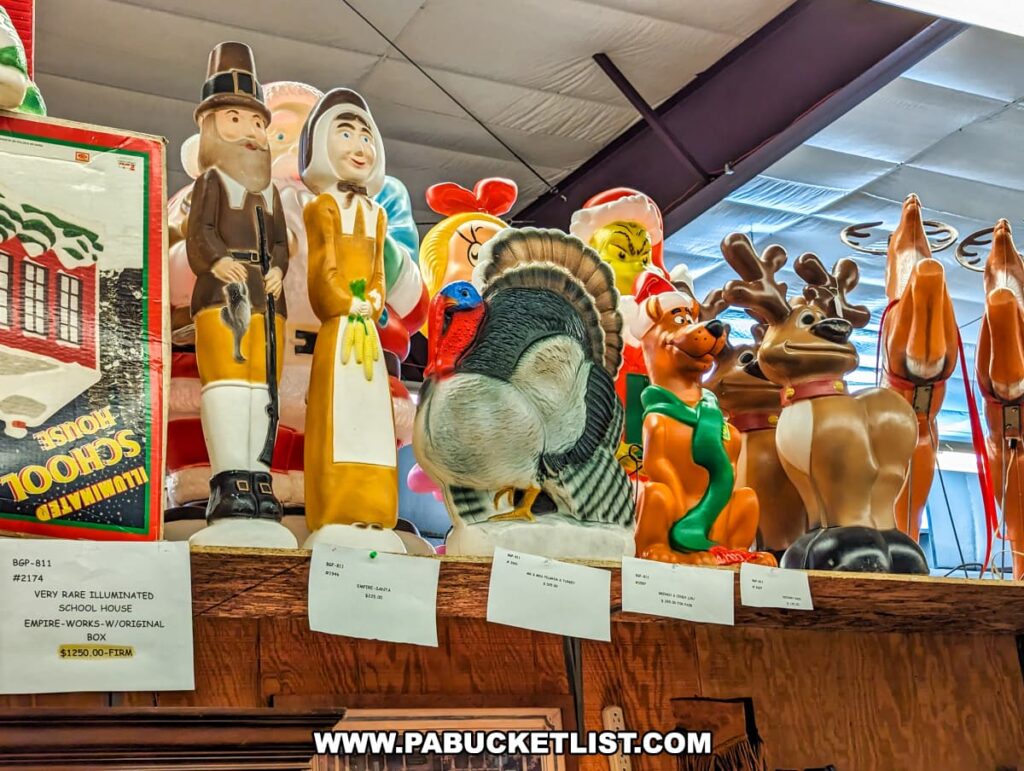 A collection of vintage blow mold holiday decorations displayed at Cackleberry Farm Antique Mall in Lancaster County, PA. The assortment includes colorful figures such as a pilgrim, a turkey, a reindeer, and other festive characters. The decorations are arranged on a high shelf, showcasing their vibrant colors and nostalgic appeal. The overhead lighting and spacious setting highlight the unique and collectible nature of these holiday items.