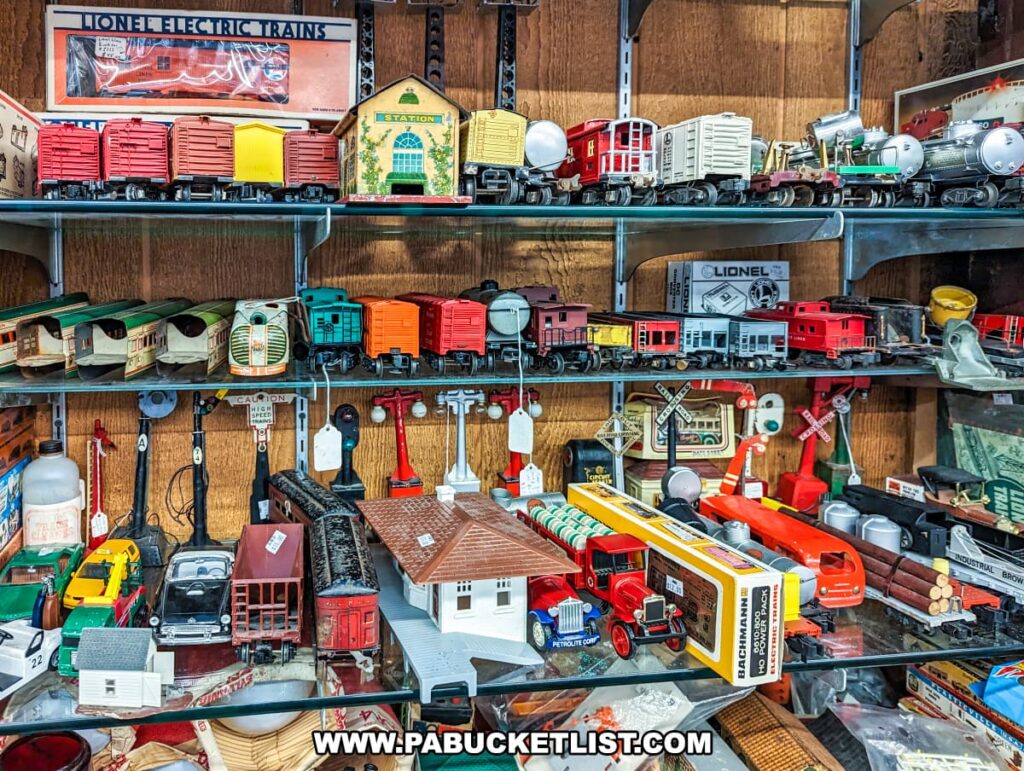 A display at Cackleberry Farm Antique Mall in Lancaster County, PA, featuring a variety of vintage model trains and accessories. The shelves are filled with colorful train cars, engines, and station models, including brands like Lionel. The collection includes detailed pieces such as railroad crossing signs, miniature buildings, and vehicles, offering a nostalgic look at model railroading. The well-organized display showcases the rich history and craftsmanship of these classic toys, appealing to collectors and train enthusiasts.