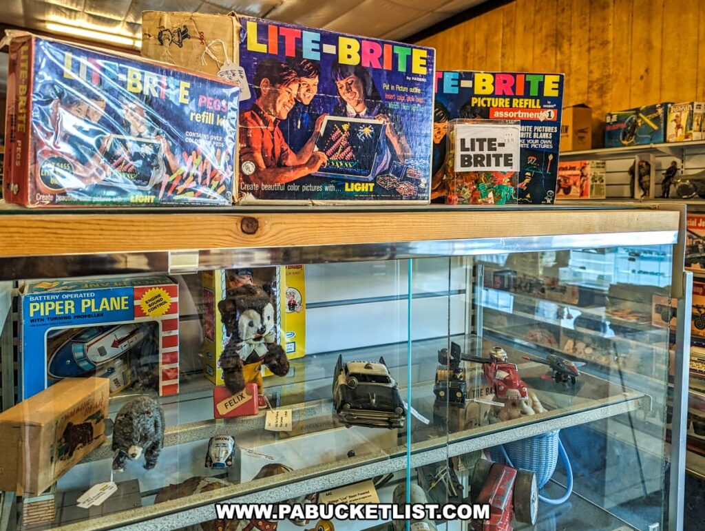 A display at Cackleberry Farm Antique Mall in Lancaster County, PA, featuring a collection of vintage toys. The top shelf showcases classic Lite-Brite sets and refill kits in their original packaging. Below, a glass case contains a variety of nostalgic toys, including a battery-operated Piper Plane, stuffed animals, and miniature vehicles. The arrangement highlights the colorful and charming designs of these timeless toys, evoking memories of childhood for visitors and collectors.