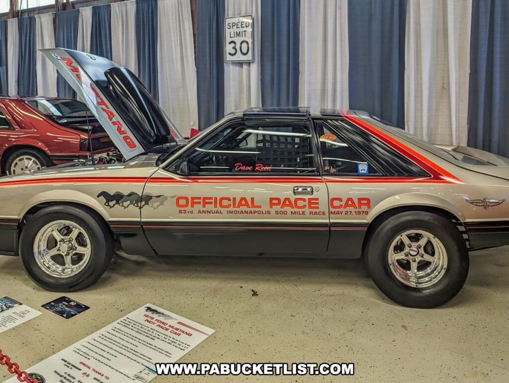 1979 Indianapolis 500 Official Pace Car, a Mustang with a distinctive silver and black paint job featuring red accents and the "Official Pace Car" designation on the side, displayed indoors at the Carlisle Events Ford Nationals car show. The car's hood is open, revealing part of the engine, with informational plaques placed nearby. Other classic Mustangs are visible in the background, along with a "Speed Limit 30" sign.