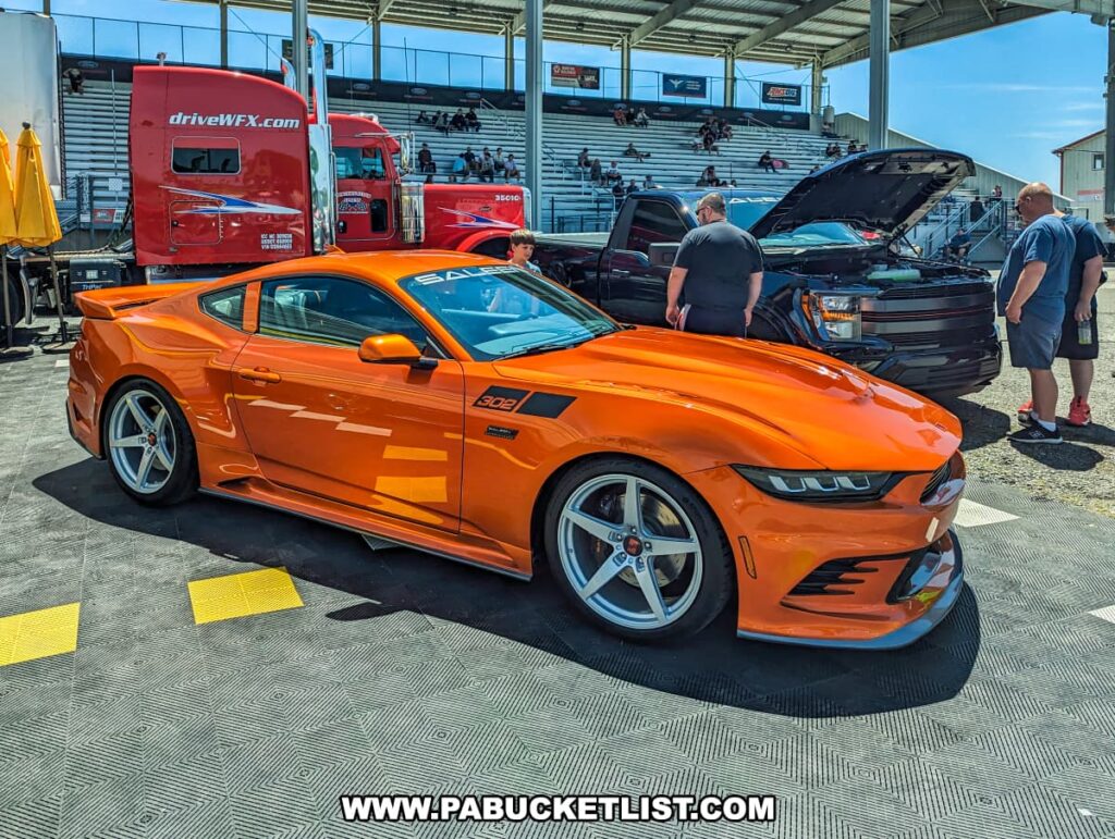 Bright orange Saleen Mustang 302 displayed at the Carlisle Events Ford Nationals car show. The car is parked on a checkered platform with its sleek, modern design prominently showcased. In the background, people are seen examining a black truck with its hood open. Red trailers with the "driveWFX.com" logo and a grandstand with seated spectators are also visible, contributing to the bustling and energetic atmosphere of the event. The sunny day and clear blue sky enhance the vibrant colors of the vehicles on display.