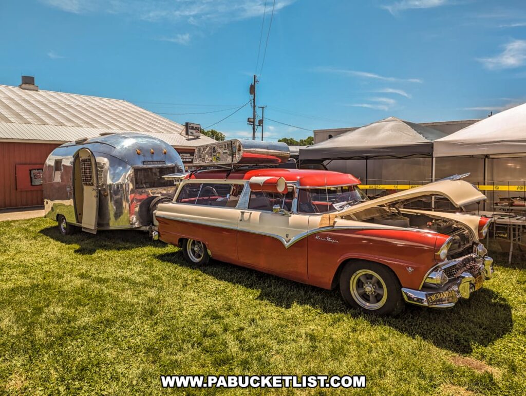 A vintage red and white Ford station wagon with its hood open, hitched to a polished Airstream camper, displayed on the grass at the Carlisle Events Ford Nationals car show. The car has a canoe mounted on its roof, adding to its classic appeal. Nearby tents and buildings are visible under a bright, sunny sky, enhancing the nostalgic atmosphere of the event.