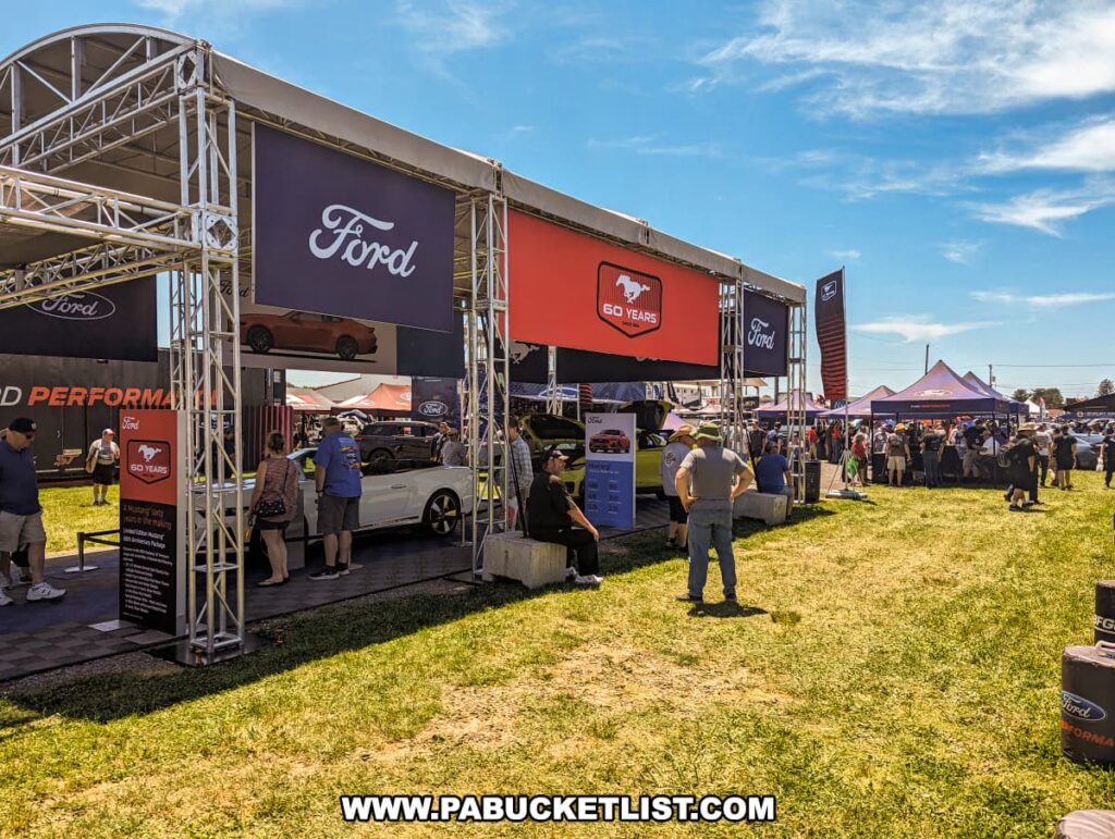 Ford display area at the Carlisle Events Ford Nationals car show, featuring banners celebrating 60 years of the Mustang. The area is bustling with attendees examining various Ford vehicles and engaging with exhibits. The setup includes a large tent structure with Ford branding and informational signs. Additional tents and displays are visible in the background under a sunny sky with scattered clouds, contributing to a vibrant and energetic atmosphere.