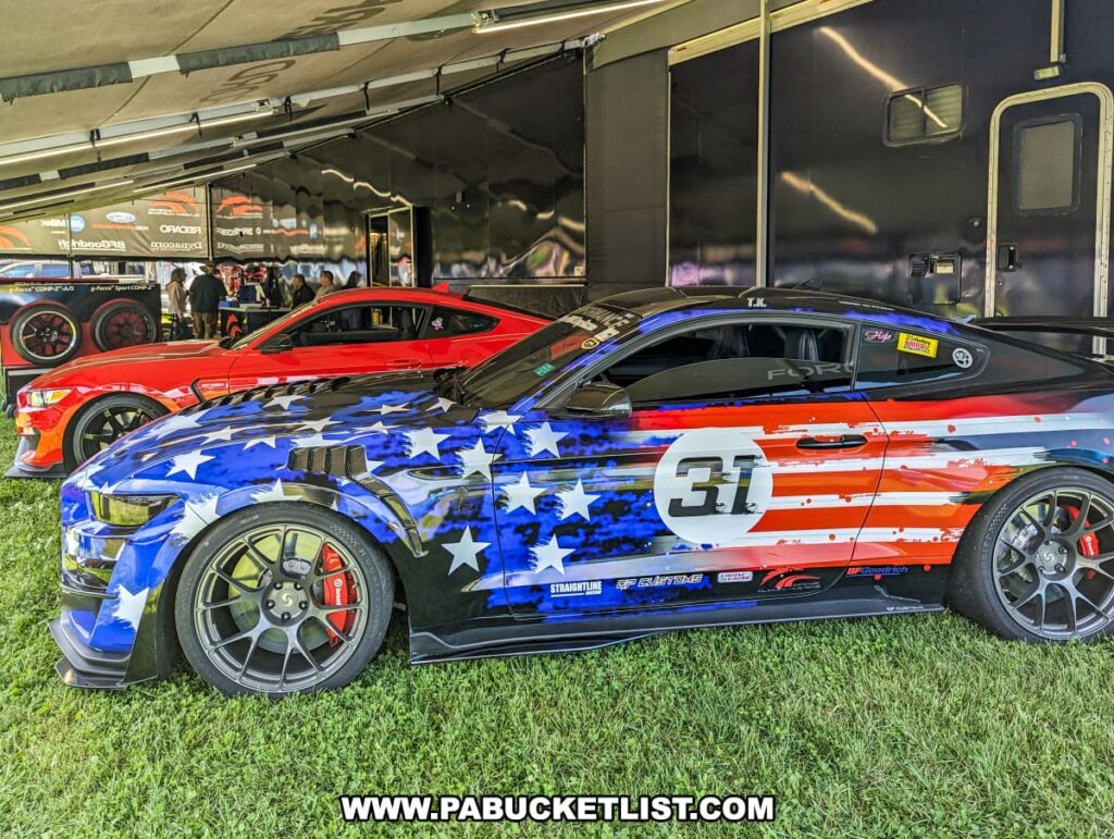 Custom Ford Mustang with a patriotic American flag paint job, displayed under a tent at the Carlisle Events Ford Nationals car show. The car features bold stars and stripes and is parked next to a bright red Mustang. The setup includes large black trailers and various equipment in the background, with people visible engaging with the displays. The grass and shade provide a comfortable environment for attendees to admire the cars on a sunny day.