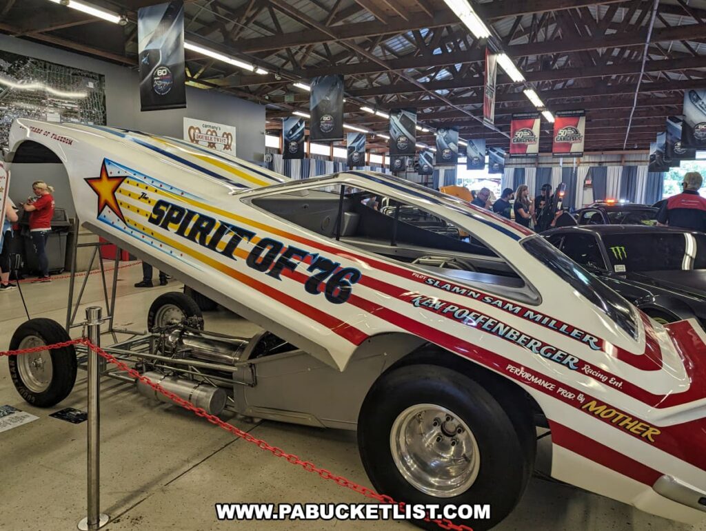 Indoor display of the "Spirit of '76" drag car at the Carlisle Events Ford Nationals car show. The car features a patriotic red, white, and blue paint job with stars and stripes, and the body is raised to showcase the chassis and engine. The surrounding area includes informational plaques and a red rope barrier. Attendees are seen in the background, examining other vehicles and engaging in conversations under a well-lit, rustic ceiling adorned with banners celebrating 60 years of Ford performance.