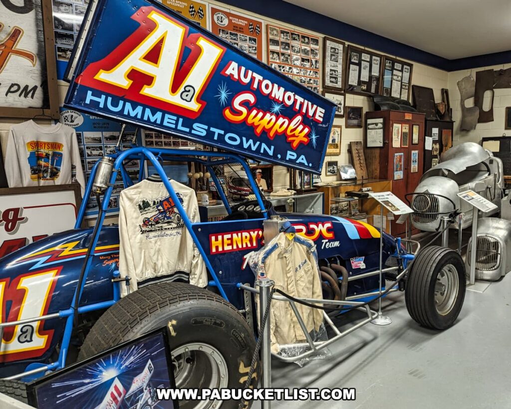 A detailed exhibit at the Eastern Museum of Motor Racing in Adams County, Pennsylvania, featuring the iconic blue A1 Automotive Supply sprint car from Hummelstown, PA. The display includes racing jackets, photos, and memorabilia, illustrating the car's history and significance in American motor racing. Behind the sprint car, various artifacts and documents are showcased, highlighting the rich heritage of motor racing in Pennsylvania and the surrounding regions. The exhibit is meticulously arranged, providing an immersive experience for visitors interested in the sport's legacy.