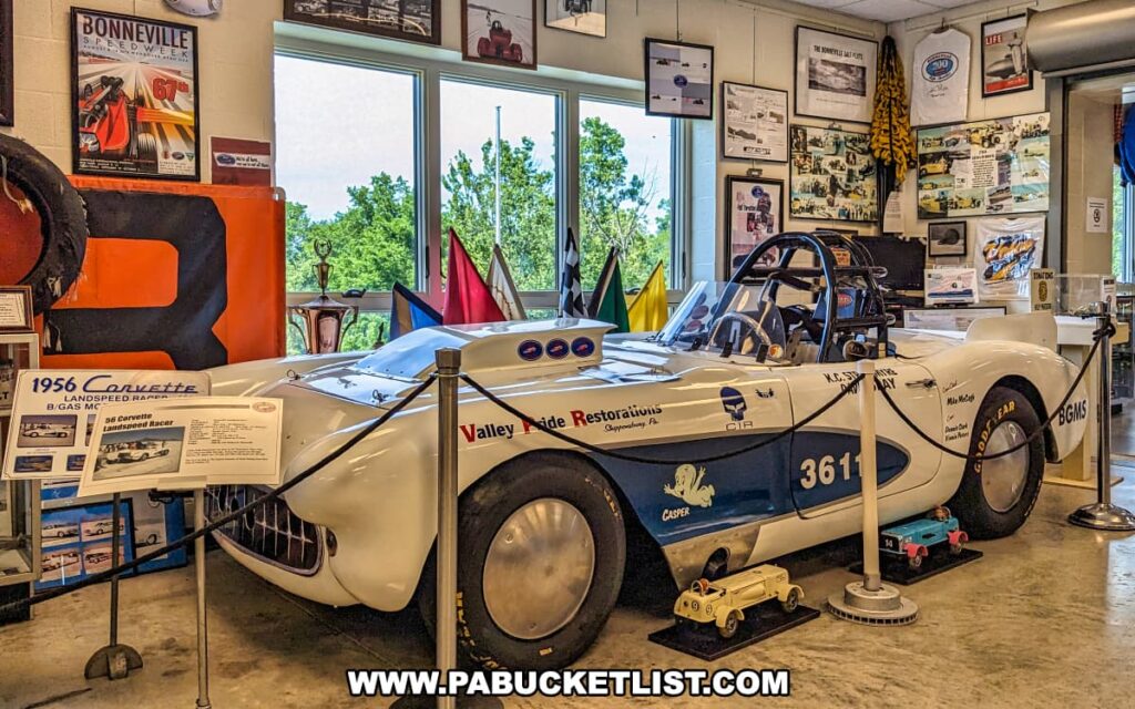 A meticulously displayed 1956 Corvette landspeed racer, restored by Valley Pride Restorations in Shippensburg, PA, at the Eastern Museum of Motor Racing in Adams County, Pennsylvania. The car, adorned with the number 361 and various sponsor decals, is surrounded by informational placards and racing memorabilia, including flags, trophies, and historical photos. The exhibit highlights the vehicle's history and achievements, particularly its participation in events like Bonneville Speedweek. The room is brightly lit with natural light from large windows, offering visitors an engaging and informative experience on the history of American motor racing.
