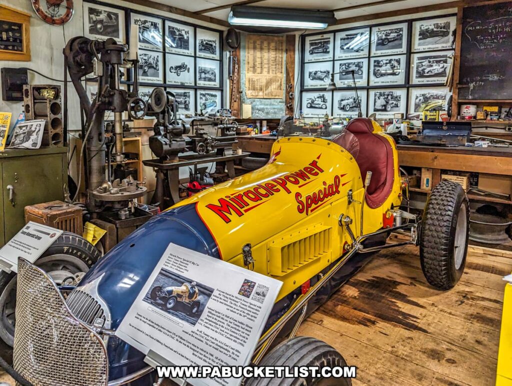 A vibrant display at the Eastern Museum of Motor Racing in Adams County, Pennsylvania, featuring the yellow "Miracle Power Special" race car driven by Tommy Hinnershitz. The exhibit includes detailed informational plaques about the car and its history. The backdrop showcases a collection of black-and-white photographs documenting various moments in motor racing history. Surrounding the car, vintage machinery and tools provide an authentic workshop atmosphere, enhancing the historical context. This exhibit aligns with the museum's mission to preserve and interpret the rich heritage of American motor racing, particularly in Pennsylvania and the surrounding regions.