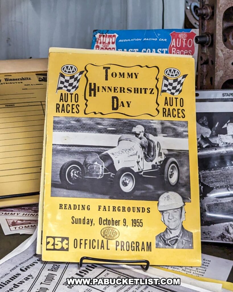 A vintage racing program from "Tommy Hinnershitz Day" held at the Reading Fairgrounds on Sunday, October 9, 1955, displayed at the Eastern Museum of Motor Racing in Adams County, Pennsylvania. The yellow program features a black-and-white photograph of a race car in action, along with an inset photo of Tommy Hinnershitz. The cover prominently displays the event details and the price of 25 cents. Surrounding the program are additional racing memorabilia and documents, reflecting the museum's mission to preserve and interpret the rich history of American motor racing, with a particular focus on Pennsylvania and the surrounding regions.