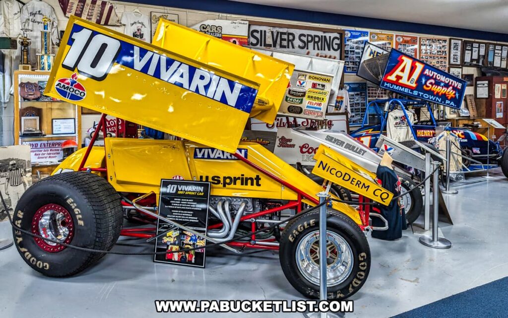 An exhibit at the Eastern Museum of Motor Racing in Adams County, Pennsylvania, featuring a bright yellow #10 Vivarin sprint car. The display includes detailed information about the car's history and significance, with signage and memorabilia from Silver Spring Speedway in the background. Adjacent to the Vivarin car is the blue A1 Automotive Supply sprint car, highlighting the variety of historic vehicles in the collection. The exhibit is surrounded by various racing artifacts, including posters, banners, and photographs, reflecting the museum's mission to preserve and interpret the rich history of American motor racing, particularly in Pennsylvania and surrounding regions.