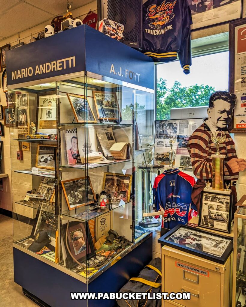 A display case at the Eastern Museum of Motor Racing in Adams County, Pennsylvania, dedicated to legendary race car drivers Mario Andretti and A.J. Foyt. The exhibit features an array of memorabilia, including photographs, trophies, race car models, and personal items. The case is surrounded by additional racing artifacts, such as jackets and framed pictures, celebrating the achievements and contributions of these iconic drivers to the sport of motor racing. The exhibit provides an in-depth look at the history and legacy of American motor racing, aligning with the museum's mission to preserve and interpret the sport's rich heritage.
