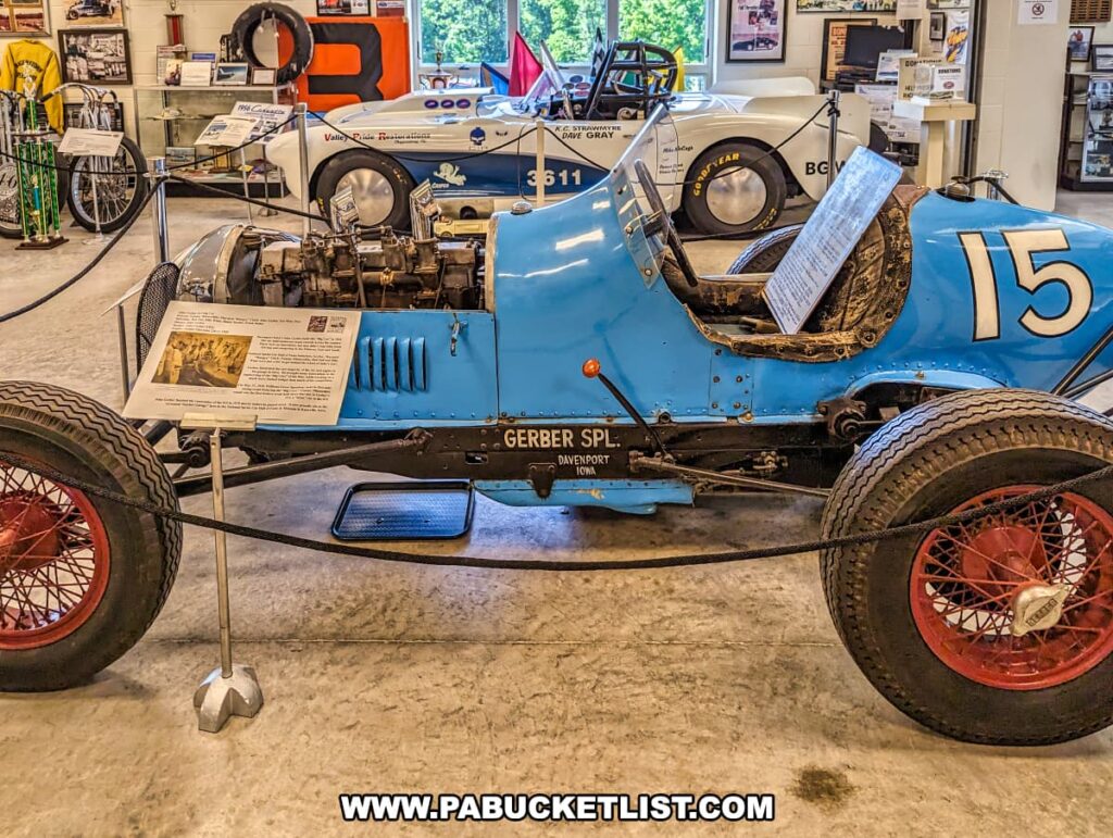 A historic blue race car, labeled "Gerber Spl." from Davenport, Iowa, displayed at the Eastern Museum of Motor Racing in Adams County, Pennsylvania. The car, marked with the number 15, is showcased with detailed informational plaques explaining its significance and history. Behind it, other racing vehicles and memorabilia, including a white 1956 Corvette landspeed racer, are visible. The exhibit is part of the museum's mission to preserve and interpret the history of American motor racing, with a focus on Pennsylvania and surrounding regions. The setting offers an engaging glimpse into the past for visitors interested in the legacy of motor racing.