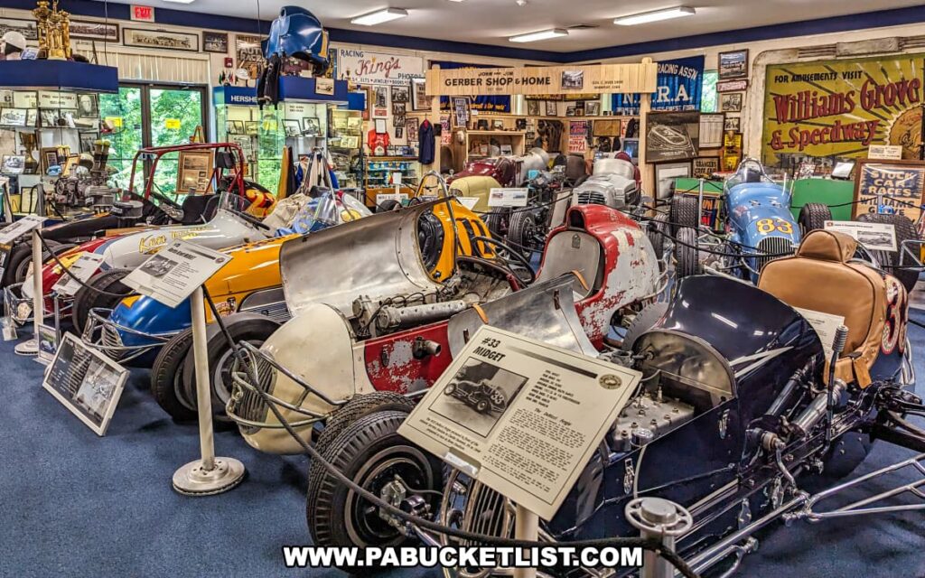 A diverse collection of historic race cars on display at the Eastern Museum of Motor Racing in Adams County, Pennsylvania. The exhibit features various vintage vehicles, including a red and white #33 Midget racer and other classic sprint cars, each accompanied by informational placards detailing their history and significance. The room is filled with memorabilia, trophies, photographs, and racing artifacts, all contributing to the museum's mission to preserve and interpret the rich history of American motor racing, with a special focus on Pennsylvania and surrounding regions. The setting is well-organized, providing an immersive and educational experience for visitors.