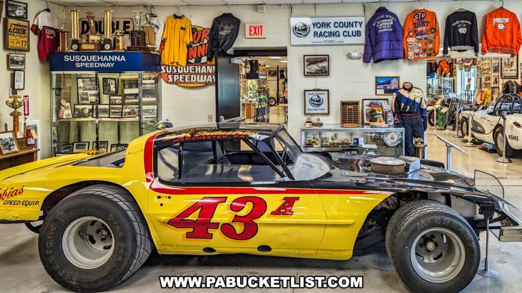 A vibrant display at the Eastern Museum of Motor Racing in Adams County, Pennsylvania, featuring a bright yellow and red #43A race car from Manchester, PA. The exhibit is enriched with an array of racing memorabilia, including trophies, jackets, photographs, and items from Susquehanna Speedway and the York County Racing Club. A mannequin dressed in vintage racing gear stands beside the car, enhancing the historical context. The well-organized setting offers an immersive experience into the rich heritage of motor racing, aligning with the museum's mission to preserve and interpret the sport's history, particularly in Pennsylvania and its neighboring regions.