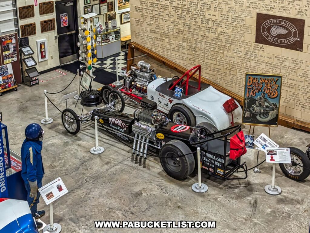 An overhead view of a detailed exhibit at the Eastern Museum of Motor Racing in Adams County, Pennsylvania, showcasing vintage drag cars. The display includes Don Garlits' Swamp Rat III dragster and other classic racing vehicles, surrounded by informational placards and memorabilia. A mannequin dressed in vintage racing gear stands nearby, enhancing the historical ambiance. The walls are adorned with plaques and posters, including a tribute to "Big Daddy" Don Garlits. The exhibit provides an immersive experience into the history of American drag racing, aligning with the museum's mission to preserve and interpret the sport's rich heritage, particularly in Pennsylvania and surrounding regions.