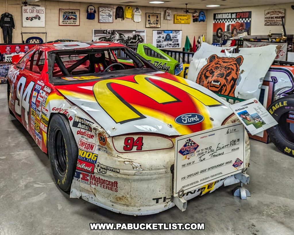 A vibrant exhibit at the Eastern Museum of Motor Racing in Adams County, Pennsylvania, featuring a vintage NASCAR race car with the number 94 and prominent McDonald's branding. The car, a Ford model, is displayed with visible wear and tear, adding authenticity to its racing history. A large ceremonial check is propped against the car's front bumper, symbolizing a racing award. Surrounding the car are additional racing artifacts, including a bear-themed car hood and various memorabilia that reflect the museum's mission to preserve and interpret the rich history of American motor racing, particularly in Pennsylvania and the surrounding regions.
