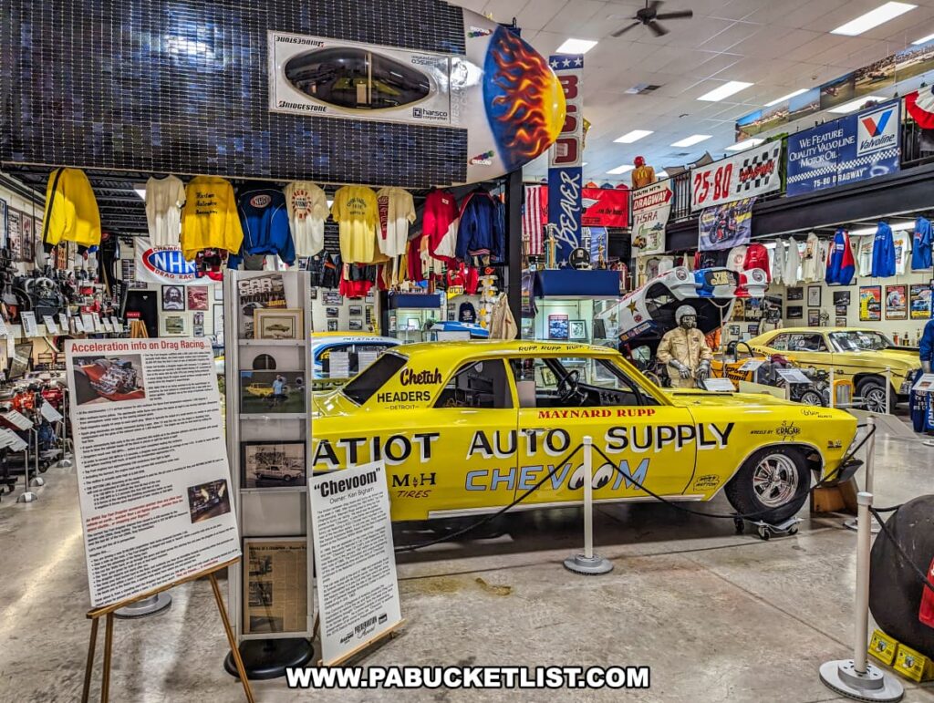 An exhibit at the Eastern Museum of Motor Racing in Adams County, Pennsylvania, showcasing a vintage yellow drag racing car labeled "Chevoom" with "Patriot Auto Supply" and "Maynard Rupp" decals. The display includes detailed informational boards about the car and the history of drag racing. The background is filled with racing memorabilia, such as jackets, banners, trophies, and photographs, creating a rich and immersive atmosphere. The upper level features additional racing items and banners, highlighting the museum's dedication to preserving and interpreting the history of American motor racing, with a special focus on Pennsylvania and surrounding regions.