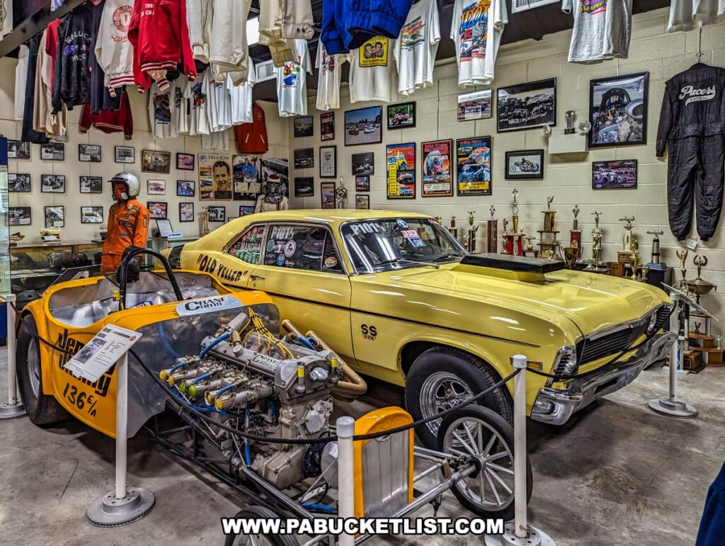 A detailed exhibit at the Eastern Museum of Motor Racing in Adams County, Pennsylvania, featuring a yellow drag racing car labeled "Old Yeller" and an orange and yellow race car with a prominently displayed engine. The background showcases a collection of racing memorabilia, including numerous trophies, framed photographs, and racing suits. The walls are adorned with historic racing shirts and jackets, adding to the immersive atmosphere. A mannequin dressed in vintage racing gear stands nearby, enhancing the exhibit's authenticity. This display aligns with the museum's mission to preserve and interpret the rich history of American motor racing, particularly in Pennsylvania and the surrounding regions.
