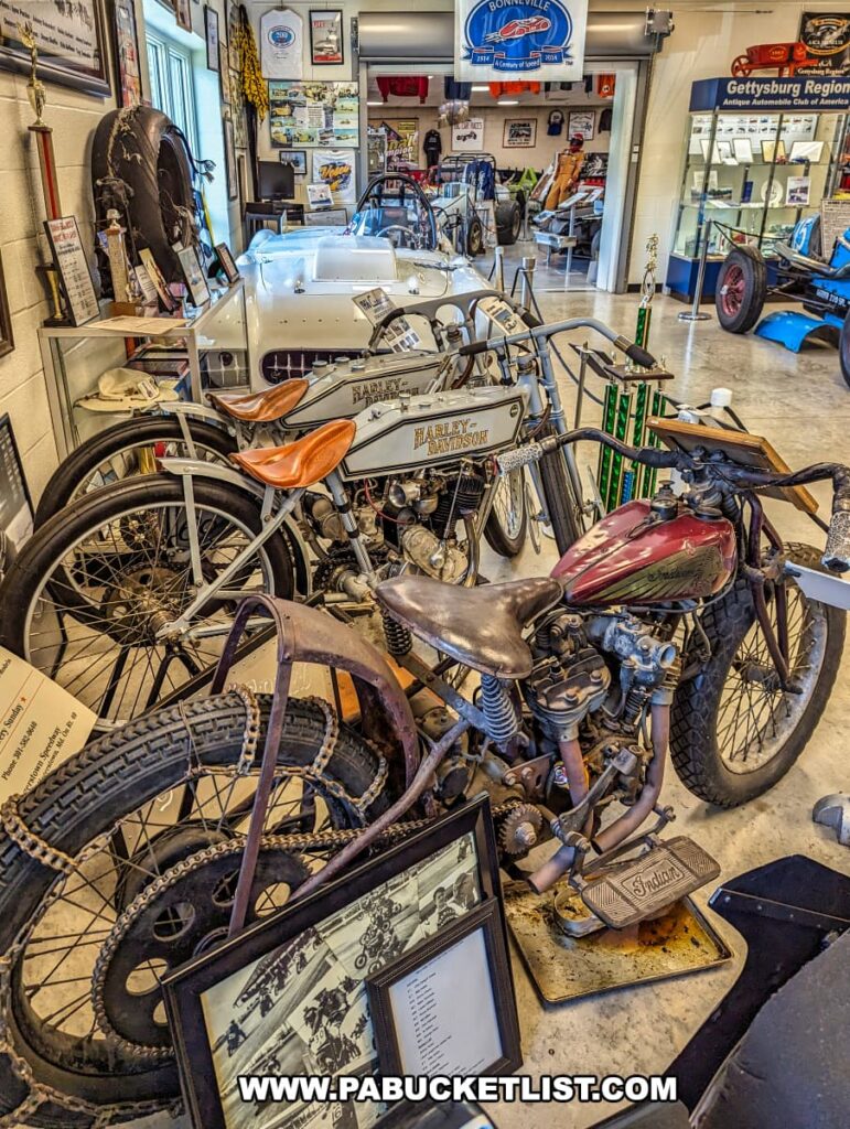 An exhibit at the Eastern Museum of Motor Racing in Adams County, Pennsylvania, featuring vintage racing motorcycles, including a Harley-Davidson and an Indian. The display showcases the bikes' aged and authentic condition, complete with leather seats and vintage tires. Surrounding the motorcycles are various racing memorabilia, including trophies, framed photographs, and informational plaques. In the background, additional historic race cars and exhibits are visible, reflecting the museum's mission to preserve and interpret the rich history of American motor racing, particularly in Pennsylvania and the surrounding regions.