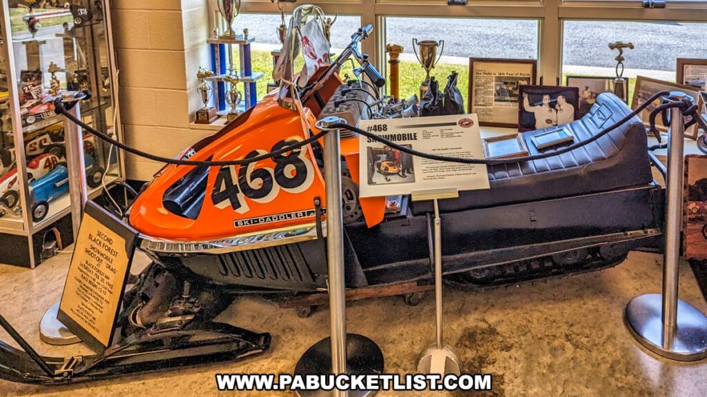 An exhibit at the Eastern Museum of Motor Racing in Adams County, Pennsylvania, featuring a vintage racing snowmobile with the number 468. The snowmobile, identified as a Ski-Daddler, is displayed with informational plaques detailing its history and significance. Surrounding the snowmobile are various racing trophies, framed photographs, and memorabilia that highlight the achievements in snowmobile racing. The exhibit is well-lit by natural light from nearby windows, providing a clear view of the items on display. This setup reflects the museum's mission to preserve and interpret the rich history of American motor racing, particularly in Pennsylvania and the surrounding regions.