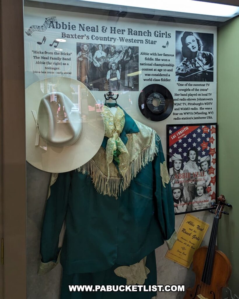 The exhibit features memorabilia of Abbie Neal & Her Ranch Girls, highlighting Baxter's Country-Western Star. The display includes a white cowboy hat, a green fringed jacket with matching scarf, and a photograph of Abbie Neal with her famous fiddle. The exhibit also mentions her early achievements, including winning a national championship at age 10 and being a TV cowgirl of the 1950s. A vinyl record and a poster of Abbie Neal & Her Ranch Girls Band are also showcased, along with historical details about her radio and TV appearances.