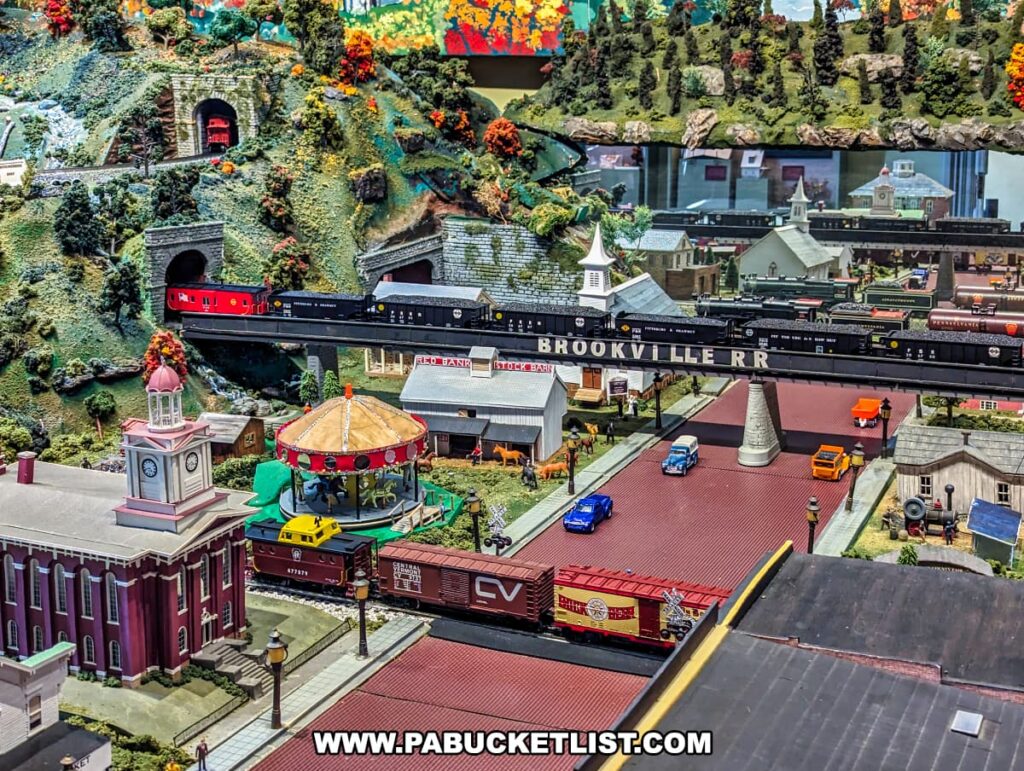A close-up view of the model railroad display at the Jefferson County History Center in Brookville, Pennsylvania. The scene features a detailed miniature town with a clock tower, a carousel, and various buildings. The "Brookville RR" train is shown crossing an elevated track above the town, with additional trains passing through tunnels in the surrounding green, mountainous landscape. The streets are lined with vintage vehicles, and the intricate details of the model create a lively and nostalgic atmosphere. The vibrant colors and meticulous craftsmanship highlight the historical and cultural significance of the exhibit.