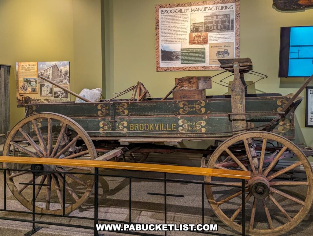 A historic wooden wagon manufactured by the Brookville Manufacturing Company, displayed at the Jefferson County History Center in Brookville, Pennsylvania. The wagon features large wooden wheels and intricate painted details, with the word "Brookville" prominently visible on its side. Behind the wagon, an informative display provides historical context about the Brookville Manufacturing Company, including its founding, operations, and eventual closure. Additional historical artifacts and exhibits are visible in the background, enhancing the historical narrative presented in the museum.