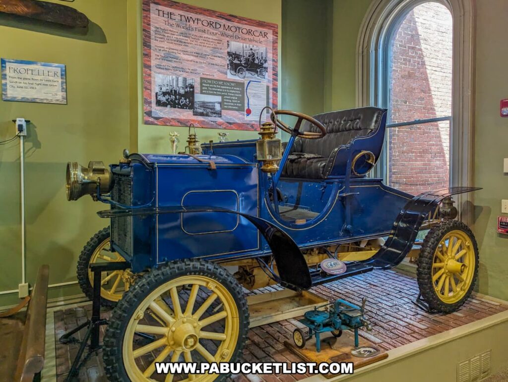 An exhibit at the Jefferson County History Center in Brookville, Pennsylvania, featuring the Twyford Motorcar. The car is showcased in a vibrant blue with yellow wheels and detailing, and it is positioned prominently in the display area. The informational panel behind the vehicle describes the Twyford Motorcar as the world's first four-wheel-drive automobile. Additional exhibits surrounding the car include a propeller from a plane flown by Lincoln Beachey and various historical artifacts. The room is well-lit, with natural light streaming through a large arched window, highlighting the significance of this pioneering vehicle in automotive history.