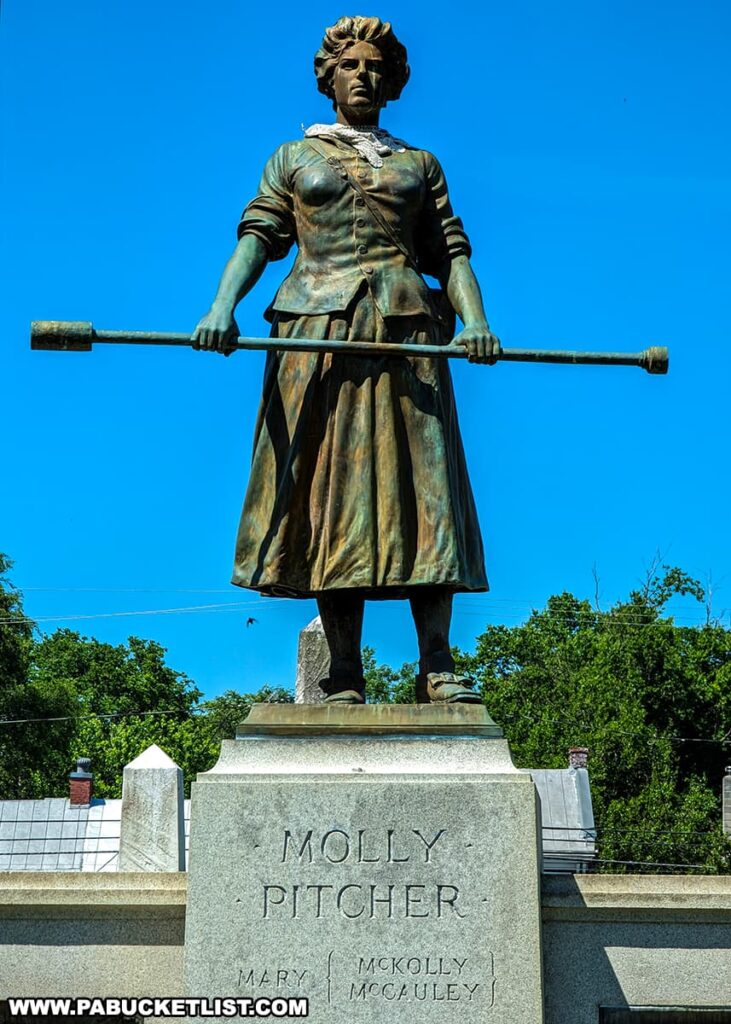 The photo shows a close-up view of the Molly Pitcher statue at her monument and gravesite in Old Cemetery, Carlisle, PA. The statue depicts Molly Pitcher standing resolutely, holding a cannon ramrod with both hands. She is dressed in period clothing, with a determined expression on her face. The pedestal beneath the statue bears the inscription "Molly Pitcher" along with "Mary McKolley McCauley." The bright blue sky and surrounding trees provide a vivid backdrop, highlighting the statue's details and the historical significance of the site.