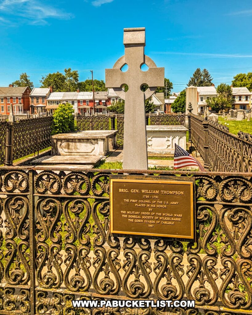 The photo shows the gravesite of Brigadier General William Thompson in Old Cemetery, Carlisle, PA, on a clear day. The site is marked by a large Celtic cross headstone and surrounded by an ornate wrought iron fence. A plaque on the fence honors General Thompson as the first Colonel of the U.S. Army and commemorates his contributions. In the background, gravestones and historical buildings are visible, contributing to the cemetery's historic ambiance. An American flag is placed near the headstone, emphasizing the patriotic significance of the site.