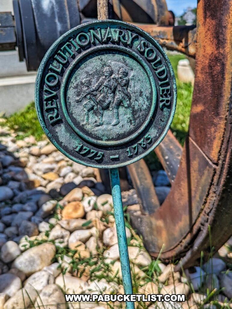 The photo shows a round bronze medallion at the base of the Molly Pitcher monument in Old Cemetery, Carlisle, PA. The medallion, attached to a metal rod, reads "Revolutionary Soldier 1775-1783" and features a raised image of two soldiers. The medallion is placed among small white rocks near the wheel of an old cannon, emphasizing its historical significance and connection to the Revolutionary War. The green patina on the bronze and the surrounding natural elements enhance the medallion's antique appearance.