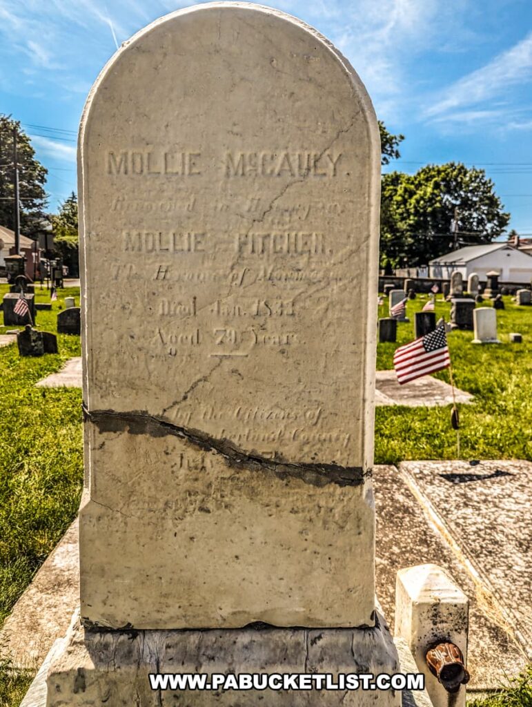 The photo shows the weathered headstone of Molly McCauley, also known as Molly Pitcher, in Old Cemetery, Carlisle, PA. The inscription reads: "Mollie McCauley, honored as Molly Pitcher, the heroine of Monmouth. Died Jan 1832, aged 79 years." The headstone has a visible crack across its middle. In the background, other gravestones and American flags are visible, highlighting the historical significance of the cemetery. The bright blue sky and lush green grass provide a serene setting for this resting place of a Revolutionary War heroine.