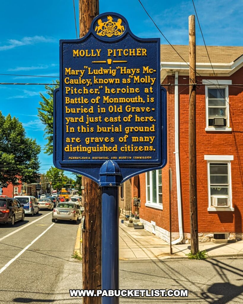 The photo shows a blue historical marker in Carlisle, PA, dedicated to Molly Pitcher. The marker reads: "Molly Pitcher. Mary Ludwig Hays McCauley, known as 'Molly Pitcher,' heroine at Battle of Monmouth, is buried in Old Graveyard just east of here. In this burial ground are graves of many distinguished citizens. Pennsylvania Historical and Museum Commission." The marker stands on a sidewalk with a street and red brick buildings in the background. The bright sunny day highlights the vibrant colors of the marker and the surrounding urban setting.
