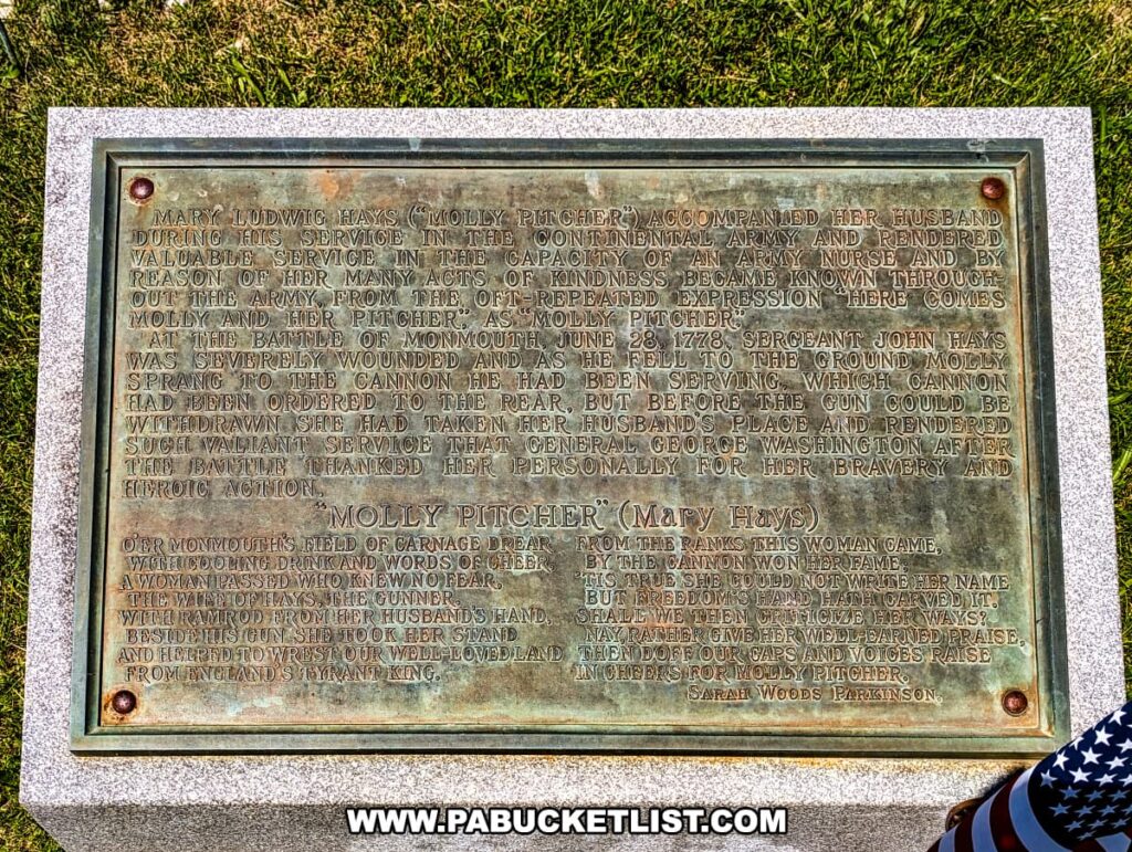 The photo shows a detailed plaque at the base of the Molly Pitcher monument in Old Cemetery, Carlisle, PA. The plaque recounts the heroic actions of Mary Ludwig Hays, known as Molly Pitcher, during the Battle of Monmouth on June 28, 1778. It describes how she took over her husband's position at the cannon after he was wounded and continued to serve bravely. The inscription also includes a poem by Sarah Woods Parkinson that honors Molly Pitcher's courage and contribution to the war effort. The plaque is set in a stone frame, with green grass surrounding it, and an American flag is visible at the edge of the photo.