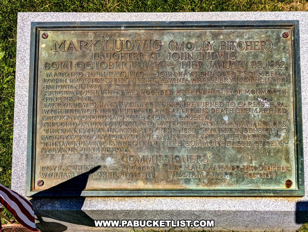 The photo shows a commemorative plaque at the base of the Molly Pitcher monument in Old Cemetery, Carlisle, PA. The plaque details the life of Mary Ludwig, known as Molly Pitcher, born on October 13, 1744, and died on January 22, 1832. It outlines her marriage to John Hays, his enlistment in the Continental Army, and her heroic actions at the Battle of Monmouth. After her husband's death, she married Sergeant George McKolly (or McCauley). The plaque also mentions the pension granted to her by the Pennsylvania Assembly for her service. The monument was erected by the Commonwealth of Pennsylvania on June 28, 1916. The grass surrounding the plaque and a partial view of an American flag add to the historical and respectful ambiance.