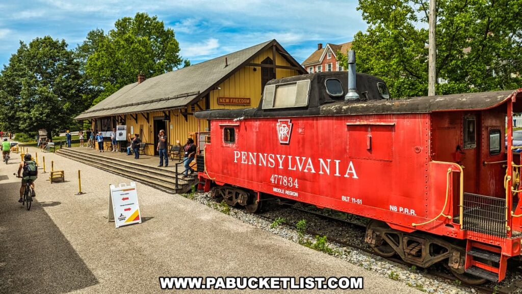 The red Pennsylvania Railroad caboose numbered 477834 is stationed next to the yellow New Freedom Station at the Northern Central Railway of York. Visitors and cyclists are seen exploring the area, enjoying the historic ambiance of the railway station. The caboose, with its vibrant color and vintage design, contrasts with the station's wooden architecture and the lush green trees in the background. The bright sky and bustling activity around the station capture the lively and nostalgic atmosphere of this historic site.