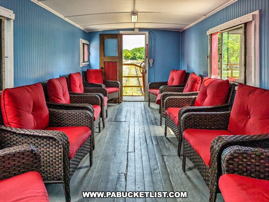 A cozy interior view of a caboose car on the Northern Central Railway of York. The car features wicker chairs with plush red cushions arranged along the blue-painted walls, creating a comfortable and inviting atmosphere. Large windows offer scenic views of the surrounding landscape, while an open door at the end of the car provides a glimpse of the tracks and greenery beyond. The wooden floor and vintage decor enhance the historical charm of the railway experience.