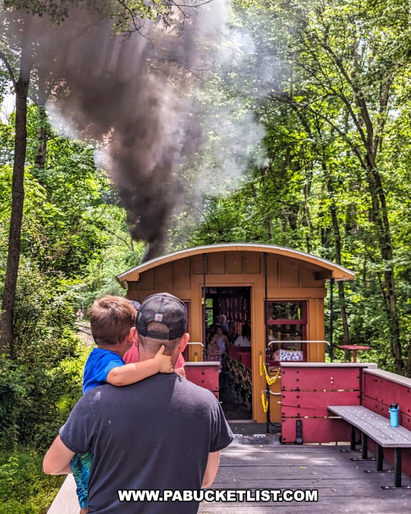A touching moment on the Northern Central Railway of York, showing a father carrying his young son while standing in an open-air gondola car. The car is painted red and features wooden benches, with other passengers visible in the background. A thick plume of black smoke rises from the steam locomotive ahead, adding to the historical ambiance. The surrounding lush forest creates a serene and picturesque setting for this memorable train ride.