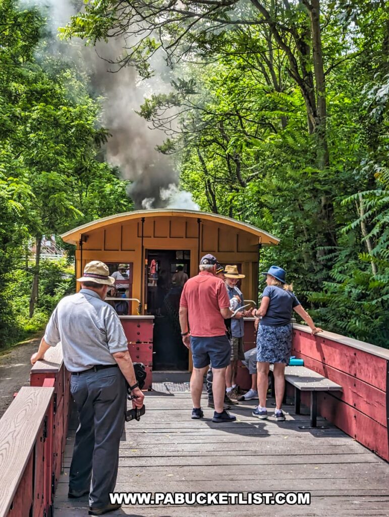 Passengers enjoying a ride in an open-air gondola car on the Northern Central Railway of York. The car is painted red and features wooden benches, with passengers standing and chatting as the train moves through a lush green forest. Thick black smoke from the steam locomotive ahead rises into the canopy, adding to the historical atmosphere. The scene captures the relaxed and social environment of the excursion, with a mix of sunlight and shade enhancing the natural beauty of the surroundings.