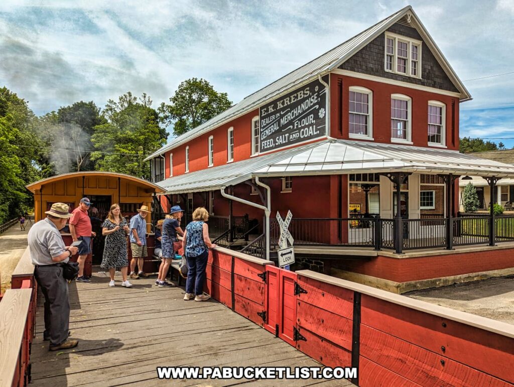 Passengers stand on an open-air gondola car at the Northern Central Railway of York, next to a historic red building labeled "E.K. Krebs, Dealer in General Merchandise, Flour, Feed, Salt, and Coal." The building features a classic design with a wraparound porch and decorative trim, adding to the historical ambiance. The scene captures the social and relaxed atmosphere of the excursion, with visitors chatting and enjoying the picturesque surroundings. The lush greenery and blue sky provide a beautiful backdrop, enhancing the charm of this historic railway experience.