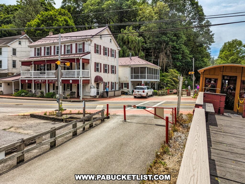 A view from the Northern Central Railway of York, looking towards a historic building with red shutters and a wraparound porch, which houses a bed and breakfast and a pub. The scene includes a railroad crossing with barriers and signs, and the tracks leading towards the station. On the right, part of the open-air gondola car is visible with passengers enjoying their ride. The lush greenery and trees surrounding the area enhance the charming and nostalgic atmosphere of this quaint town and its historic railway.