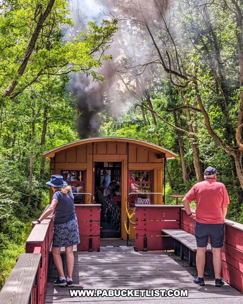 Passengers standing in an open-air gondola car on the Northern Central Railway of York. The car is painted red and has wooden benches for seating. Thick black smoke billows from the steam locomotive at the front of the train, creating a dramatic contrast with the lush green trees surrounding the railway. The dense forest and bright sunlight create a picturesque and immersive experience for the riders, highlighting the scenic and historical charm of the excursion.