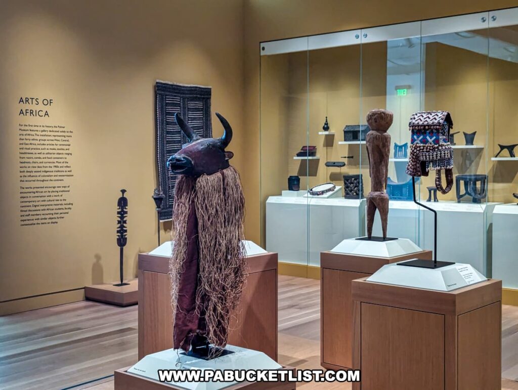 A display of African art at the Palmer Museum of Art in Centre County, PA. The exhibit features various artifacts, including a bull-headed mask with long raffia strands, a wooden statue, and a beaded garment on individual pedestals. The background shows additional African art pieces in a glass case and a wall plaque titled "Arts of Africa" providing context for the exhibition. The gallery is well-lit with a warm color scheme, enhancing the intricate details of the artifacts.