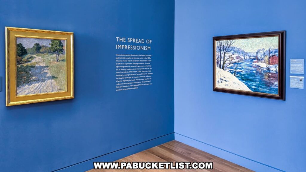 An exhibit on Impressionism at the Palmer Museum of Art in Centre County, PA. The display features two framed paintings on a blue wall. The left painting, surrounded by a gold frame, depicts a sunlit pathway through a grassy field with trees. The right painting, in a dark frame, shows a snowy riverside scene with buildings and smoke in the background. Between the paintings, a text panel titled "The Spread of Impressionism" provides historical context about the art movement and its impact in the United States. The wooden floor and minimalist design of the gallery enhance the focus on the artworks.