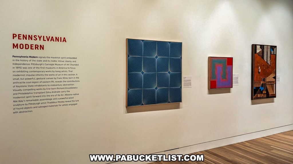 A display titled "Pennsylvania Modern" at the Palmer Museum of Art in Centre County, PA. The exhibit features three modern art pieces on a white wall. On the left, a blue geometric painting with a grid-like pattern. In the center, a smaller abstract painting with overlapping pink, red, and blue shapes. On the right, a mixed-media piece incorporating various textures and colors. The wall text explains the significance of modern art in Pennsylvania, highlighting the state's history of fostering contemporary works and the contributions of artists such as Franz Kline, Richard Anuszkiewicz, Edna Andrade, and Abe Ajay. The wooden floor and minimalist presentation enhance the focus on the artworks.